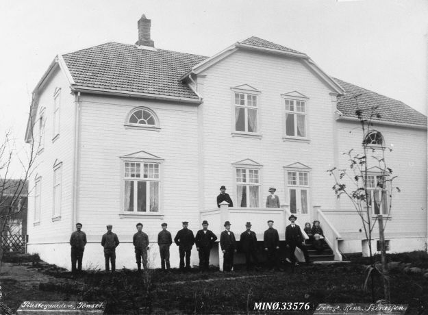 sort hvitt bilde av prestegården med dresskledde menn foran fasaden