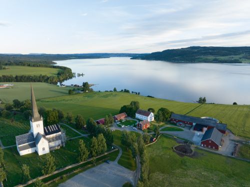 dronebilde av kirke og gårdsbruk med bygninger som ligger til innsjø