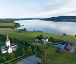 dronebilde av kirke og gårdsbruk med bygninger som ligger til innsjø