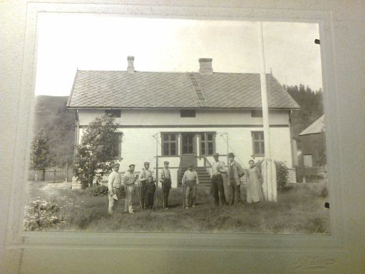 Sort hvitt gammelt bilde der mange arbeidsfolk er avfotografert  foran huset .