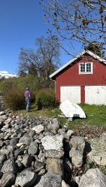 et stort, rødmalt naust ligger i fjordkanten. Foran er det svaberg og store steiner, i bakgrunnen fjell med snø på toppene, blå himmelr. En mann betrakter naustet