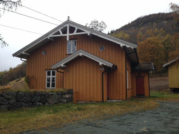 gavlside på okergul bygning med busker og vei på siden