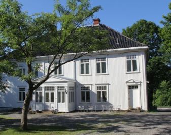 Hvitt trehus omgitt av grønne trær på tunet.
