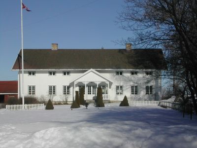 Stort hvitt trehus i vinterskrud med flaggstang og hage i forgrunnen