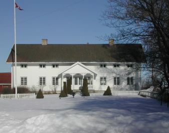 Stort hvitt trehus i vinterskrud med flaggstang og hage i forgrunnen