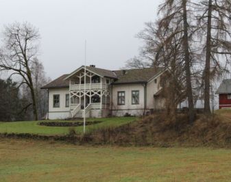 Beigemalt trehus med flaggstand og hage i forgrunnen