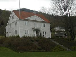 Kvitmalt bygning med flaggstang og hage i forgrunnen