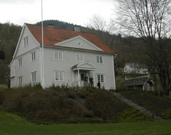 Kvitmalt bygning med flaggstang og hage i forgrunnen