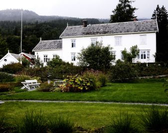Meldal prestebolig. Hvitt trehus omgitt av grønn mark og blomsterbusker.
