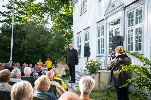 Magne Olav Brevik står foran et hvitt hus og taler til en gruppe folk